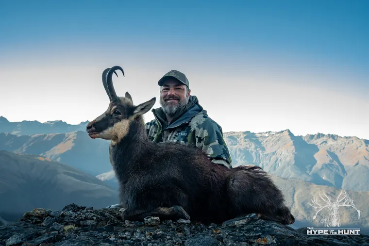 Hunter with a Himalayan tahr above layered New Zealand mountain ranges